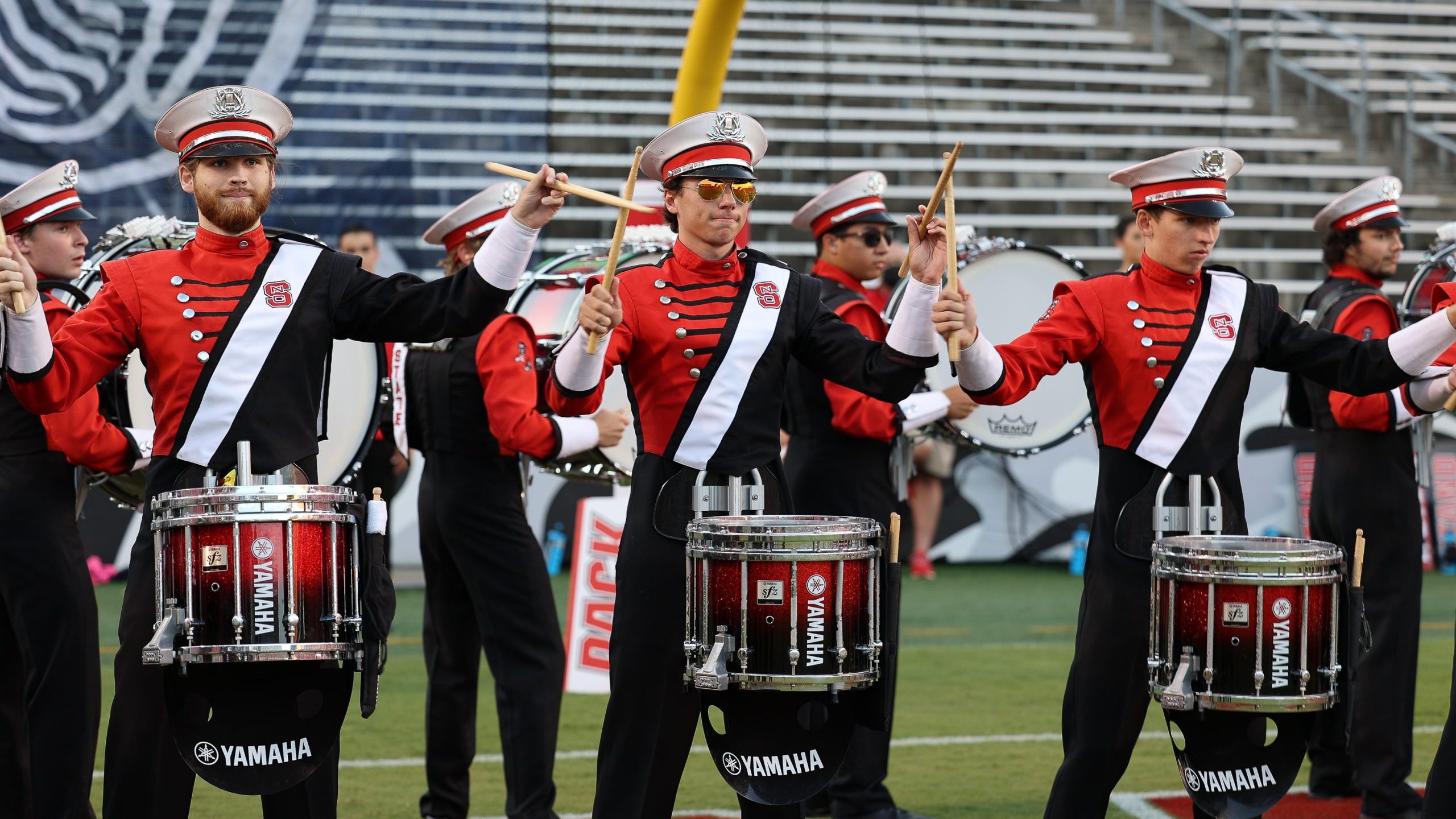 Students in marching band uniforms play drums on the field at Carter-Finley stadium.