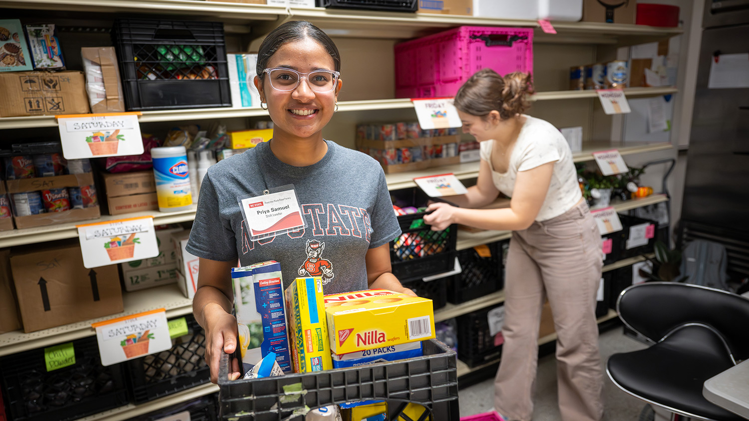 A student volunteer carries a crate of food donations while another student stocks a shelf at Feed the Pack food pantry.